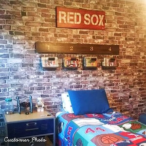 Puede incluir: Un dormitorio con un acento de pared de ladrillo, una cama con un edredón de temática deportiva y un estante con cuatro cestas etiquetadas del 1 al 4. Un letrero sobre el estante dice "RED SOX".