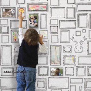 May include: A white wall with a black and white wallpaper design of various picture frames. A child is standing in front of the wall, reaching up to touch the wallpaper.