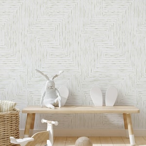 Op de afbeelding: Een witte houten bank met een lichtgrijze pluche konijn erop. De muur achter de bank heeft een geometrisch patroon van dunne gouden lijnen.