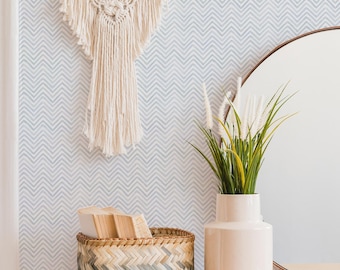 May include: A cream-colored macrame wall hanging with fringe, a rectangular woven basket with a green and tan ombre pattern containing books, and a smooth cream ceramic vase holding green grass-like plants on a wooden desk.