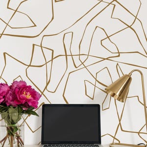 Puede incluir: Un escritorio blanco con una lámpara de escritorio dorada, un ordenador portátil plateado y un jarrón de vidrio transparente con flores rosas. La pared detrás del escritorio tiene un patrón abstracto blanco y dorado.