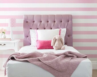 May include: A tufted mauve headboard on a bed with white sheets, a mauve quilted blanket, two white pillows with pink trim, a small rectangular hot pink pillow, and a plush rabbit toy. The wall features horizontal pink and white stripes.