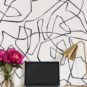 Puede incluir: Un escritorio blanco con un papel tapiz de patrón abstracto en blanco y negro, un ordenador portátil plateado, una lámpara de escritorio dorada y un jarrón de vidrio transparente con flores rosas.