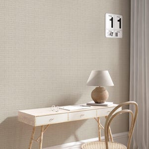 May include: A light wood desk with three drawers and a white top. A wicker chair sits in front of the desk. A lamp with a white shade and a brown base sits on the desk. A calendar with the date "11" and the day "SAT" is on the wall. The wall is covered in a beige wallpaper with a subtle pattern.
