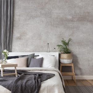 May include: A bedroom interior with a white bed, adorned with grey and neutral cushions and a dark grey throw. A small wooden tray holds a vase of white flowers and a cup. A potted plant sits on a wooden stool. A grey curtain is on the left.