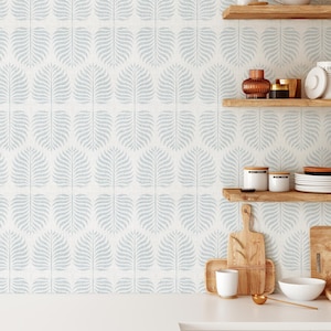 May include: A kitchen with white cabinets and a white worktop. The backsplash is a light blue wallpaper with a repeating pattern of stylised leaves. There are two wooden shelves with various kitchen items, including a teapot, mugs, jars, and plates.