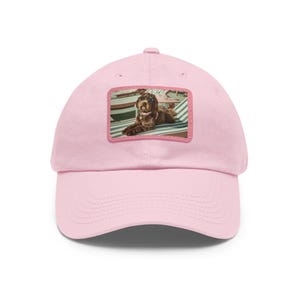Könnte beinhalten: Hellrosa Baseballkappe mit gebogenem Schirm. Die Kappe hat einen rechteckigen Aufnäher mit einem Bild eines braunen Hundes, der auf einer gestreiften Oberfläche liegt. Der Hund ist das zentrale Element des Aufnähers.