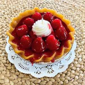 May include: A red strawberry pie candle with a white whipped cream topping and a single candle in the center. The pie is on a white doily on a woven brown surface.