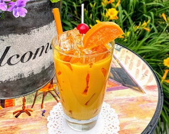 May include: A glass of orange juice with ice and a slice of orange, garnished with a cherry. The drink is on a white doily on a table with a painted design.