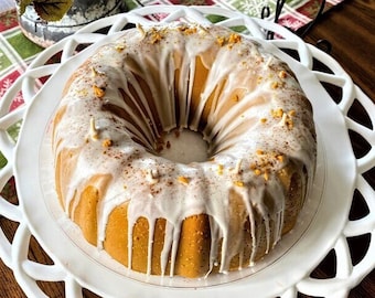 May include: A white cake with a white glaze and a sprinkle of cinnamon on top. The cake is on a white cake stand with a decorative edge.