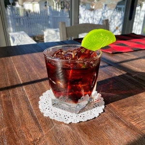 May include: A glass of dark red liquid with ice cubes and a lime wedge on the rim, sitting on a white doily on a wooden table.