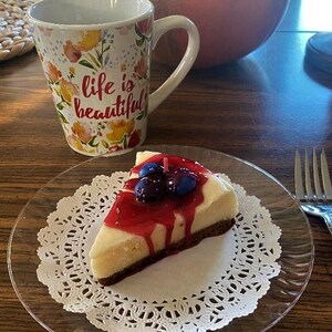 May include: A slice of cheesecake topped with blueberries and a red sauce on a white doily on a clear glass plate. The plate is on a wooden table with a mug that says "life is beautiful" on it.