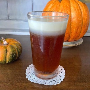 May include: A glass candle with a brown and white layered design, sitting on a white doily on a wooden table. A small orange and green pumpkin is in the background.