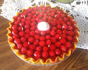 May include: A red cherry pie candle with a white frosting topping. The candle is in a yellow pie crust and has a realistic appearance.