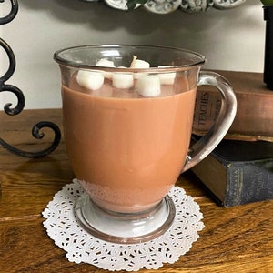Puede incluir: Una vela en forma de taza de vidrio con aroma a chocolate marr&oacute;n y malvaviscos blancos en la parte superior. La vela est&aacute; sobre una servilleta de encaje blanca.