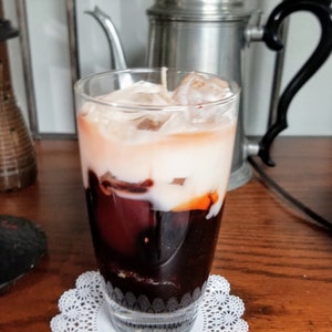 May include: A glass of iced coffee with a layer of cream on top. The coffee is dark brown and the cream is white. The glass is sitting on a white doily.
