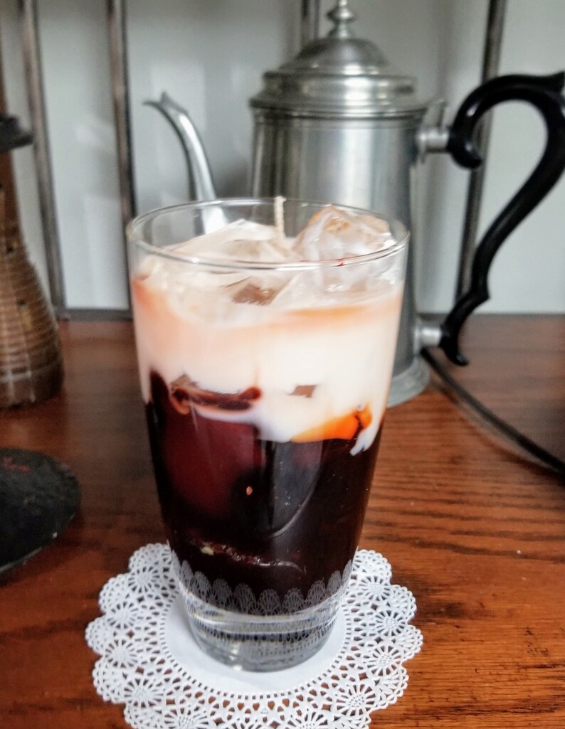 May include: A glass of iced coffee with a layer of cream on top. The coffee is dark brown and the cream is white. The glass is sitting on a white doily.