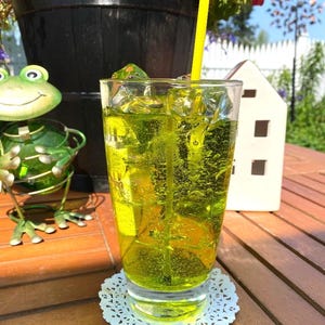 May include: A tall glass filled with a vibrant green beverage and ice cubes, with a yellow straw. The drink sits on a white lace doily on a wooden table. A decorative frog and a small white house are in the background.