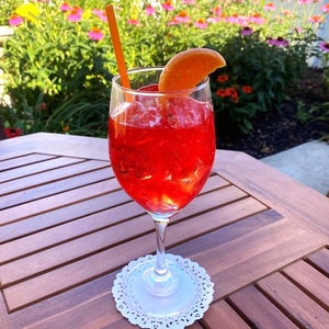 Puede incluir: Un vaso de cóctel rojo con hielo y una rodaja de naranja como guarnición, sobre un mantel individual blanco, sobre una mesa de madera con un fondo borroso de flores rosas.