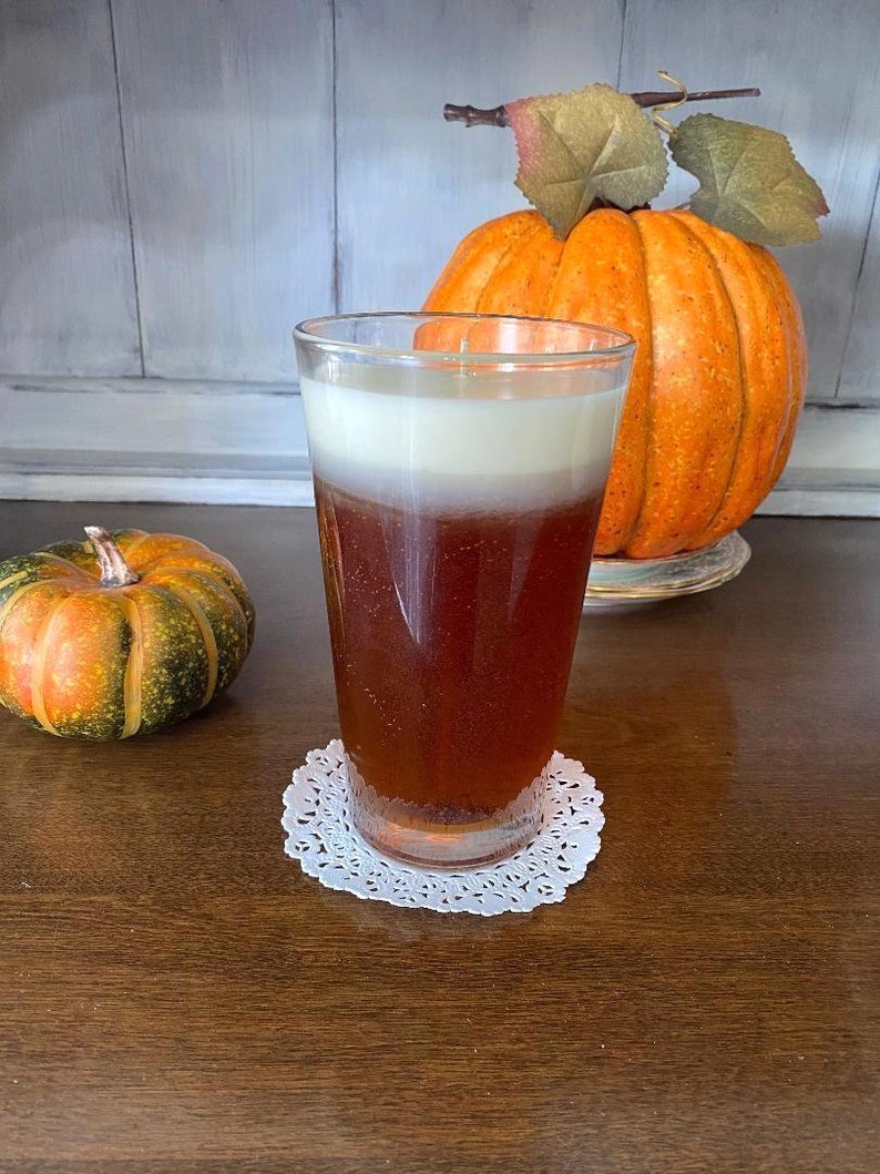 May include: A glass candle with a brown and white layered design, sitting on a white doily on a wooden table. A small orange and green pumpkin is in the background.