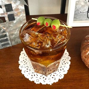May include: A glass of amber-colored liquid with ice cubes, garnished with three green olives on a toothpick. The drink is on a white doily.