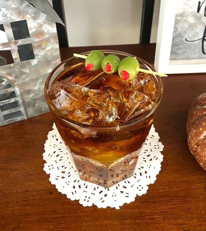 May include: A glass of amber-colored liquid with ice cubes, garnished with three green olives on a toothpick. The drink is on a white doily.
