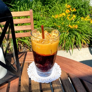 May include: A glass of iced coffee with a brown straw, topped with a layer of cream and ice cubes. The coffee is in a clear glass and is sitting on a white doily on a wooden table.
