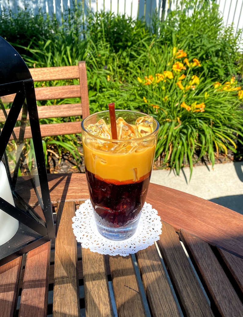 May include: A glass of iced coffee with a brown straw, topped with a layer of cream and ice cubes. The coffee is in a clear glass and is sitting on a white doily on a wooden table.