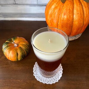 May include: A glass candle jar with a white and brown layered candle. The candle is sitting on a white doily on a wooden surface. There is a small orange and green pumpkin in the foreground and a large orange pumpkin in the background.