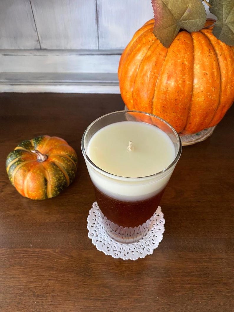 May include: A glass candle jar with a white and brown layered candle. The candle is sitting on a white doily on a wooden surface. There is a small orange and green pumpkin in the foreground and a large orange pumpkin in the background.