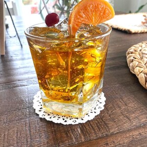 May include: A glass of amber-colored cocktail with ice cubes, a cherry on a stick, and an orange slice garnish. The drink is in a rocks glass and is sitting on a white doily on a wooden table.