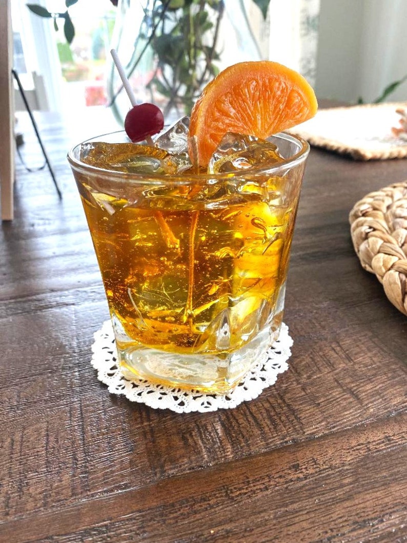 May include: A glass of amber-colored cocktail with ice cubes, a cherry on a stick, and an orange slice garnish. The drink is in a rocks glass and is sitting on a white doily on a wooden table.