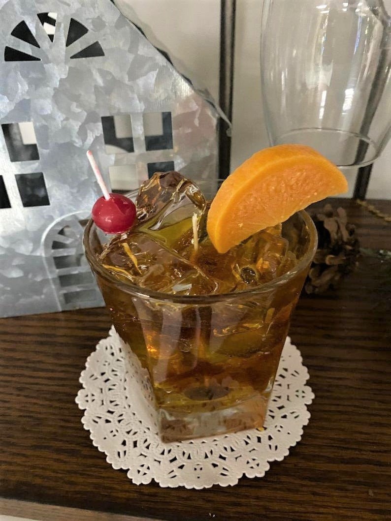 May include: A glass of whiskey with ice and an orange slice garnish. The glass is on a white doily and a red cherry is on a toothpick.