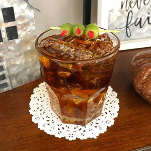 May include: A glass of brown liquid with ice cubes and three green olives on a toothpick. The glass is sitting on a white doily.