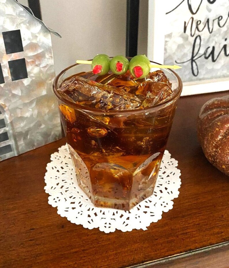 May include: A glass of brown liquid with ice cubes and three green olives on a toothpick. The glass is sitting on a white doily.