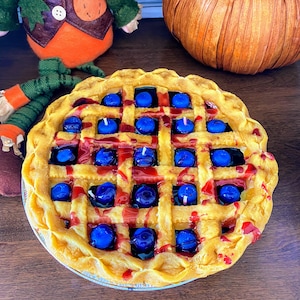 Puede incluir: Una vela decorativa con forma de tarta con un diseño de corteza de rejilla. La vela presenta "bayas" de cera azul y acentos de "jarabe" rojo, creando una apariencia de postre realista. La vela está sobre un plato plateado.