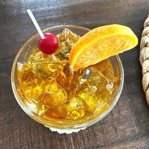May include: A glass filled with ice and a yellow alcoholic beverage, garnished with an orange slice and a maraschino cherry.