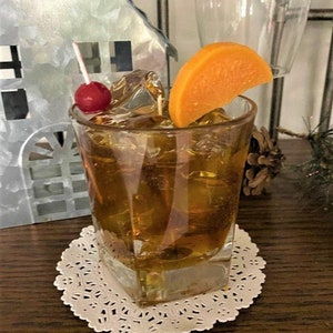 May include: A square glass filled with ice and a brown liquid, garnished with an orange slice and a maraschino cherry on a toothpick. The glass is sitting on a white doily.