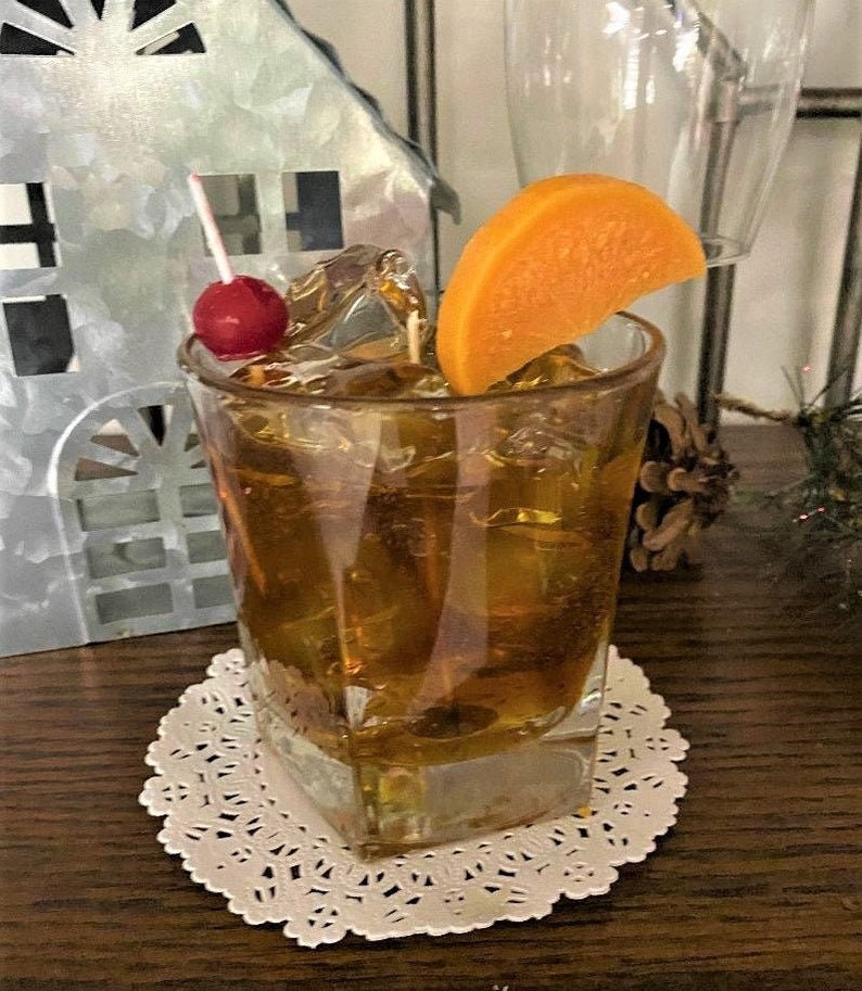 May include: A square glass filled with ice and a brown liquid, garnished with an orange slice and a maraschino cherry on a toothpick. The glass is sitting on a white doily.