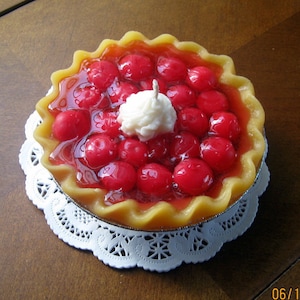 May include: A cherry pie candle with a white frosting topping. The candle is on a white doily.