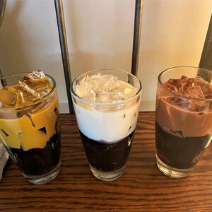 May include: Three tall glasses filled with iced coffee drinks. The glass on the left has a layer of caramel-colored coffee on top of a layer of dark coffee and ice. The middle glass has a layer of white milk on top of a layer of dark coffee and ice. The glass on the right has a layer of chocolate-colored coffee on top of a layer of dark coffee and ice.