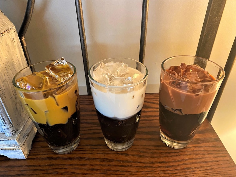 May include: Three tall glasses filled with iced coffee drinks. The glass on the left has a layer of caramel-colored coffee on top of a layer of dark coffee and ice. The middle glass has a layer of white milk on top of a layer of dark coffee and ice. The glass on the right has a layer of chocolate-colored coffee on top of a layer of dark coffee and ice.