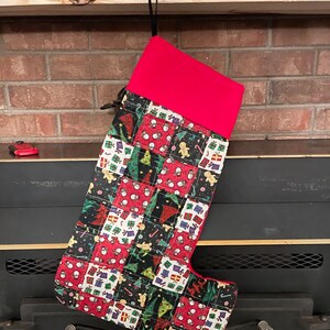May include: A red and black Christmas stocking with a patchwork design featuring presents, snowmen, and Christmas trees. The stocking is hanging from a hook above a fireplace.