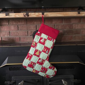 May include: A red and green quilted Christmas stocking with a patchwork design featuring small Christmas trees. The stocking is hanging from a hook on a wooden shelf above a black fireplace.