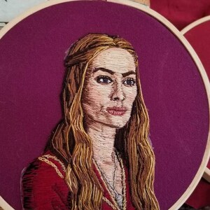 Peut inclure: Portrait brodé d'une femme aux longs cheveux blonds. Elle porte une robe rouge avec des accents dorés. La broderie est réalisée sur un tissu violet et est encadrée dans un cercle en bois.