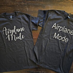 May include: Two dark gray t-shirts with the text "Airplane Mode" printed in white on the front. One shirt has the text in a cursive font, while the other shirt has the text in a block font.