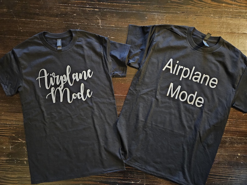 May include: Two dark gray t-shirts with the text "Airplane Mode" printed in white on the front. One shirt has the text in a cursive font, while the other shirt has the text in a block font.