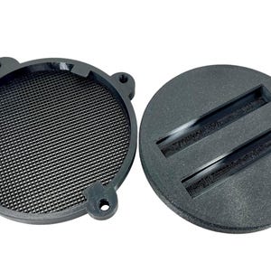 May include: Two-piece black speaker component set. The first piece is a circular frame with a mesh screen. The second piece is a solid, round base with two recessed channels. The set is designed for audio equipment.