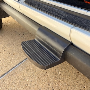 May include: A black textured side step accessory attached to a tubular rock rail on a white vehicle. The step features a ribbed surface for traction and is positioned below a door sill plate with the word GLADIATOR embossed in black plastic.