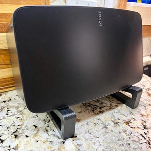 May include: A black Sonos speaker with the brand name printed on the front, resting on a black stand. The speaker has a rounded rectangular shape and a textured surface. The stand is made of a matte black material.